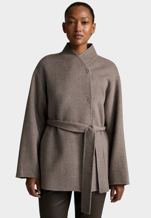 Donna che indossa un cappotto avvolgente in lana color taupe con maniche ampie e una cintura abbinata annodata in vita, in piedi contro uno sfondo neutro.