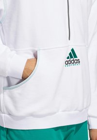 Felpa bianca con accenti turchesi, colletto con zip, polsini a coste e tasca frontale. Presenta il logo ricamato di Adidas all'orlo.