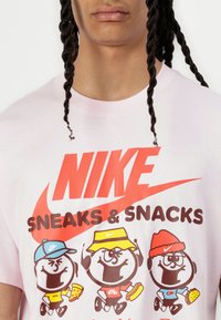 Lyserød t-shirt med et grafikprint med tre tegneseriekarakterer, der hver bærer farverige hatte, holder madvarer, og teksten "NIKE SNEAKS & SNACKS."