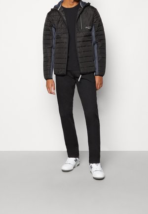 Homme portant une doudoune noire avec des panneaux latéraux gris, un pantalon noir, un t-shirt noir, et des baskets blanches avec des logos noirs, debout devant un fond blanc.