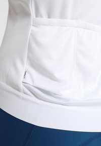 Camiseta deportiva blanca sin mangas con una textura suave, un diseño ajustado y un detalle de bolsillo en la parte posterior. Combinada con leggings azules.
