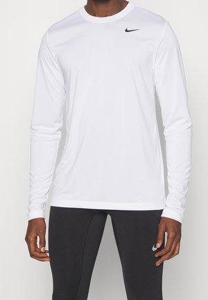 Mężczyzna ubrany w prostą białą sportową koszulkę z długim rękawem oraz czarne legginsy sportowe z logo Nike na piersi koszulki i na nodze.