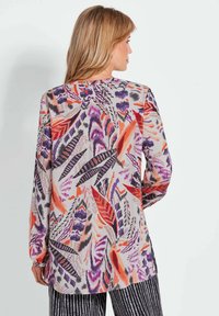 Blouse à manches longues colorée présentant un motif de feuilles vibrantes en rouge, violet et noir sur un fond clair avec des fentes sur les côtés.