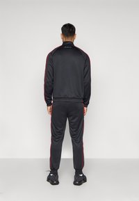 Training suit negru cu dungi roșii pe lateral, realizat dintr-un material neted. Include o jachetă cu fermoar și guler în picioare, cu manșete și talie elasticizate.