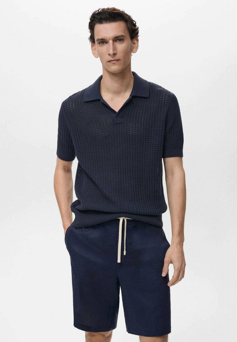 Polo in maglia texturizzata blu marina con maniche corte, con colletto e piccolo bottone frontale. Indossato con pantaloni corti blu scuri e dettaglio con cordoncino.