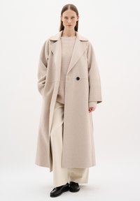 Cappotto oversize beige in lana con ampi risvolti, un bottone nero solo e tasche laterali, abbinato a un maglione leggero e pantaloni beige larghi.