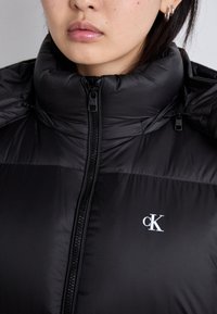 Črna puhovka z visokim ovratnikom, z zadrgo spredaj in belim logotipom "CK" na prsih. Gladka, sijoča tekstura.