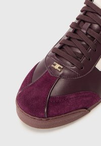Burgundy sneakers featuring a leather and suede upper, textured finish, gold logo detail, and flat laces. Rubber sole with contrasting accents.