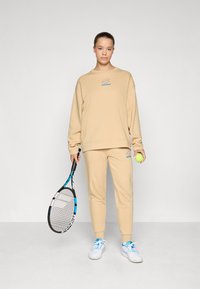 Beige sweatshirt and matching joggers with the "Lotto" logo, holding a tennis racket and ball, featuring blue accents and a relaxed fit.