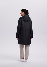 Mujer de espaldas, vestida con un impermeable negro con capucha, pantalones beige con puños elásticos y zapatillas blancas, sobre un fondo liso.