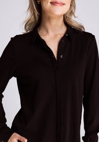 Schwarzes Langarmhemd mit Button-Down-Kragen, das eine glatte Stoffstruktur und einen dezenten Glanz aufweist. Enthält Knöpfe auf der Knopfleiste.