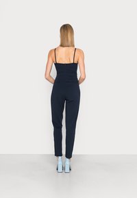 Mörkblå jumpsuit med tunna axelband, åtsittande liv och avsmalnande ben; har en dragkedja på ryggen. Kombineras med ljusblå klackskor.