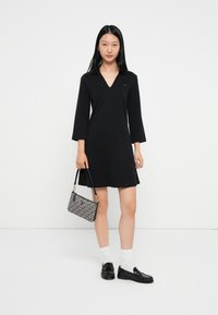 Czarna sukienka z dekoltem w serek i rękawami do trzech kwart, zestawiona z wzorzystą torebką i czarnymi mokasynami. Modelka nosi białe skarpetki.