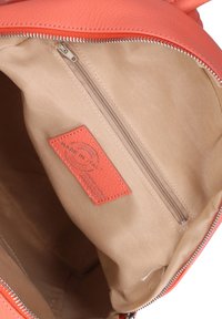 Interno di uno zaino in pelle di un beige chiaro con un'etichetta arancione. Presenta una tasca con zipper e una superficie liscia. Chiusura con zipper nella parte superiore.