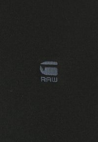 Svart tyg med en mjuk textur som har en liten, ljusgrå broderad logotyp som står "RAW." Enkelt och diskret design.