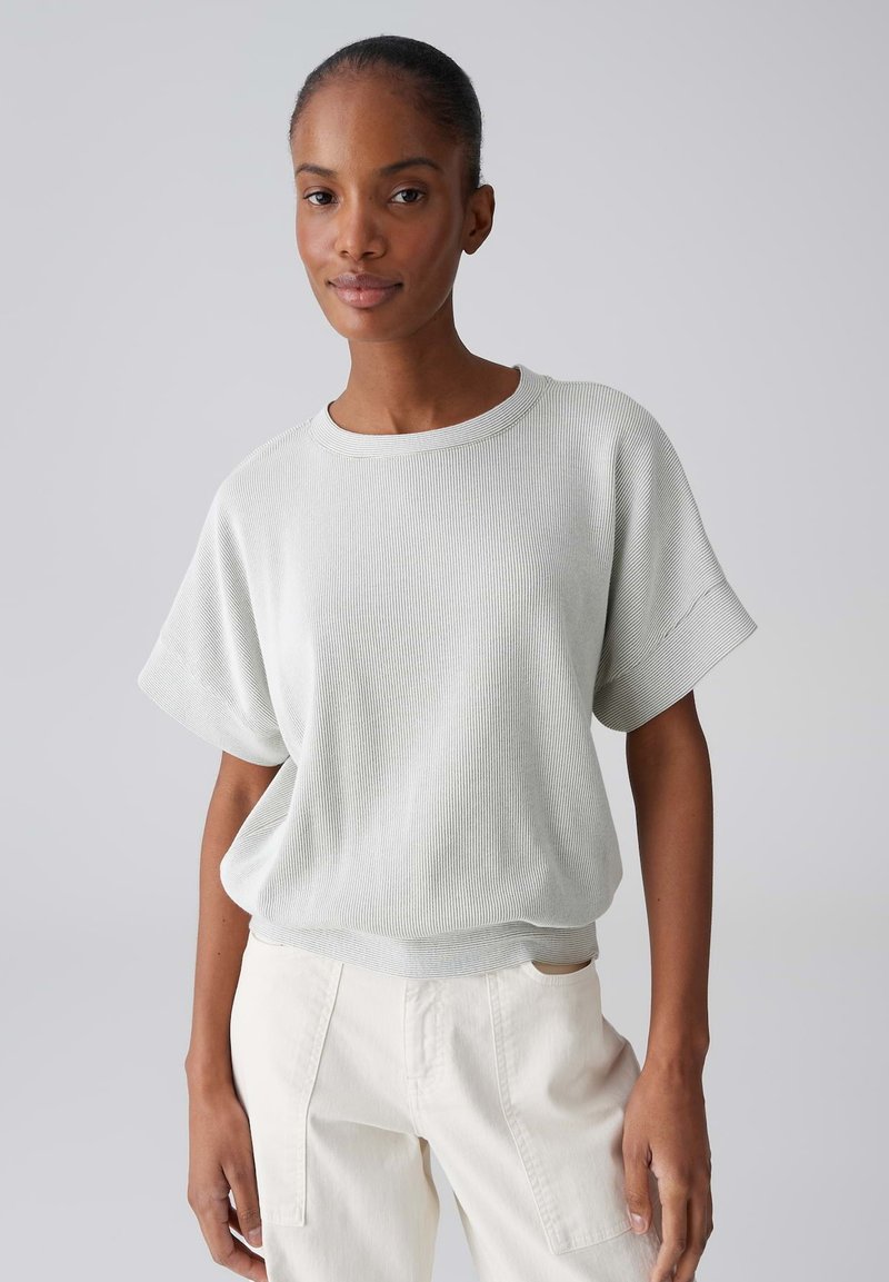 Vrouw die een losse lichtgrijze ribgebreide korte mouwen shirt en crèmekleurige broek draagt, staand tegen een effen lichtgrijze achtergrond.