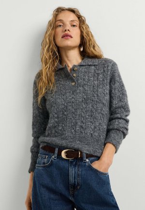 Donna con i capelli lunghi e ricci che indossa un maglione grigio lavorato a maglia con bottoni e jeans blu con cintura marrone.