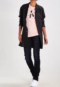 Svart, lätt jacka med knäppning framtill, bärs över en ljusrosa t-shirt med tryck och mörka jeans, kompletterad med vita sneakers.