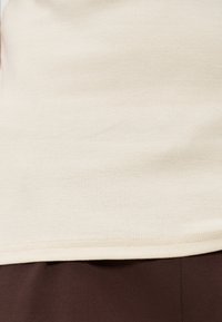 Εγγύς λήψη τελειώματος ριγωτού πουκάμισου μπεζ χρώματος πάνω από παντελόνι από σκούρο καφέ ύφασμα.