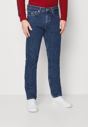 Mann trägt blaue Jeans, weiße Sneakers und ein rotes langärmeliges Hemd, steht vor einem schlichten hellen Hintergrund.