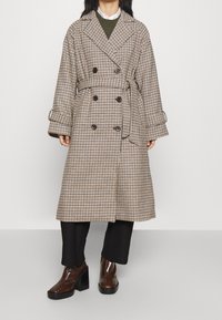 Trench coat lungo a quadri in beige e marrone, doppiopetto con bottoni scuri, vita cintata e maniche larghe, abbinato a pantaloni neri e stivali lucidi.