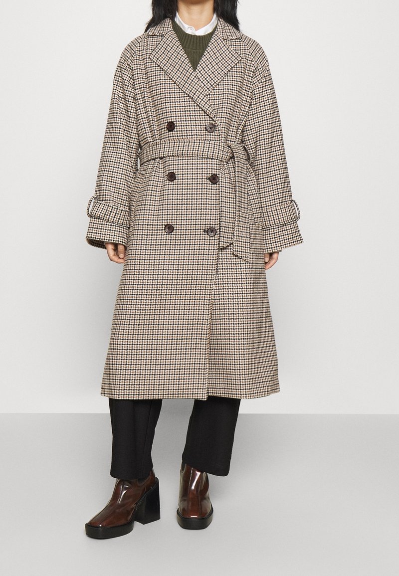 Trench coat lungo a quadri in beige e marrone, doppiopetto con bottoni scuri, vita cintata e maniche larghe, abbinato a pantaloni neri e stivali lucidi.