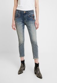 Jeans de mezclilla azul recortados con un lavado desgastado, bolsillos delanteros acentuados y costuras decorativas. Combinados con botas de tobillo de estampado de serpiente negro.