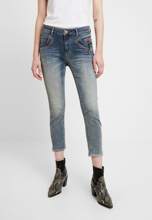Jeans de mezclilla azul recortados con un lavado desgastado, bolsillos delanteros acentuados y costuras decorativas. Combinados con botas de tobillo de estampado de serpiente negro.