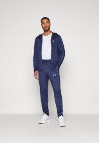 Marineblå idrettsdrakt som har en hettegenser med glidelås og lyseblå aksenter, samt matchende bukser med elastisk midje. Hvite joggesko.