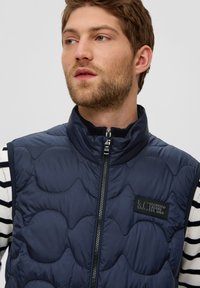 Navyblauer, gesteppter Weste mit hohem Kragen und Frontreißverschluss. Verfügt über ein Logo-Patch auf der linken Seite, das über einem gestreiften Langarmshirt getragen wird.