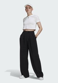 Baltas trumpas marškinėlis su juodu logotipu, aukšto liemens juodi plisuoti kelnių, ir balti sportbačiai. Modelis vilki baltą kepurę.