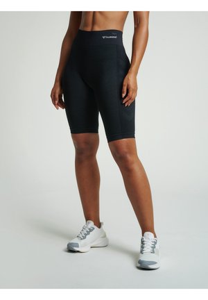 Kvinde iført sorte atletikshorts med høj talje og hvide træningssko står mod en ensfarvet baggrund med hænderne afslappet ned langs siderne.
