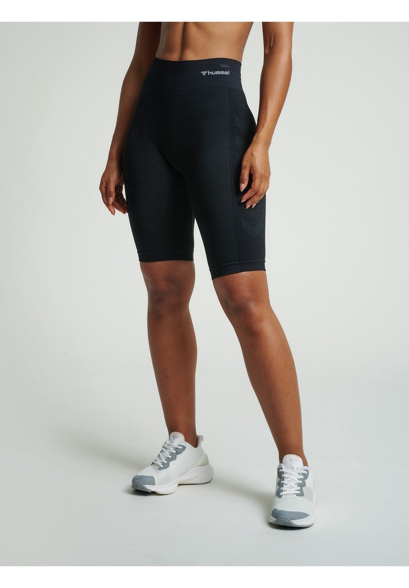 Femme portant un short de sport noir taille haute et des baskets blanches, debout devant un fond uni, les mains détendues le long du corps.