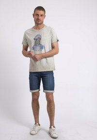 T-shirt gris à manches courtes avec un imprimé graphique d'un marin barbu portant des lunettes de soleil. Associé à un short en denim bleu et des baskets blanches.