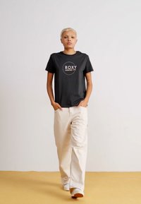 Juodas trumpomis rankovėmis marškinėliai su balta "ROXY" logotipu, derinami su laisvomis, šviesiai smėlio spalvos kelnėmis. Paprastas dizainas, švelni tekstūra, laisvas fasonas.