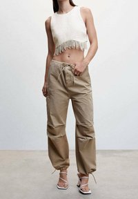 Femme portant un crop top sans manches blanc avec des franges et un pantalon cargo beige à cordon, assorti de sandales à talons blanches à lanières.