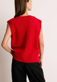 Maglione smanicato rosso in maglia con finitura testurizzata, scollatura rotonda e orlo a costine, abbinato a pantaloni in denim neri.