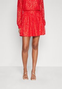 Femme portant une mini-jupe en dentelle rouge avec des manches longues et des sandales ouvertes à talons dorés, se tenant contre un fond clair.