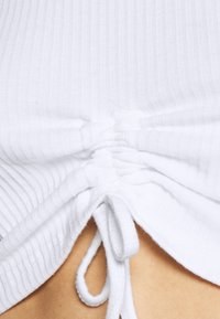 Vit ribbad topp med samla sidor som säkras med dragskor. Slät struktur, kroppsnära design och subtila finishdetaljer.