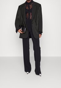 Blazer noir oversized avec détails en franges, blouse en dentelle noire transparente, pantalon noir sur mesure et sac à main noir. Escarpins pointus avec accents argentés.