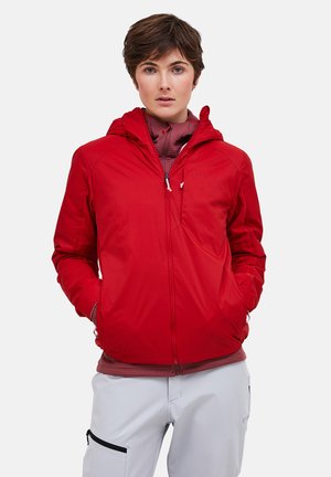 Person iført en rød hættejakke over en mørkerød lynlåstrøje, med lysegrå bukser med en sort lynlåslomme på venstre lår.