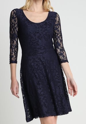 Vrouw in een marineblauwe kanten jurk tot op de knie met driekwart doorzichtige mouwen en een ronde hals, staand tegen een effen achtergrond.