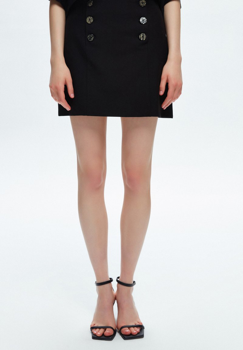 Mini falda negra con dos botones plateados en la parte delantera, combinada con tacones negros de tiras. Textura de tela suave.