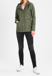 Olivengrøn, vandtæt jakke med hætte, frontknapper og sidelommer. Kombineret med sorte jeans og hvide sneakers.