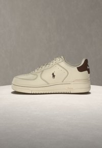 Λευκό sneaker χαμηλού τύπου με ανοιχτό καφέ ταμπελάκι στη φτέρνα και το λογότυπο Polo στο πλάι, τοποθετημένο σε ουδέτερη επιφάνεια με μονόχρωμο φόντο.