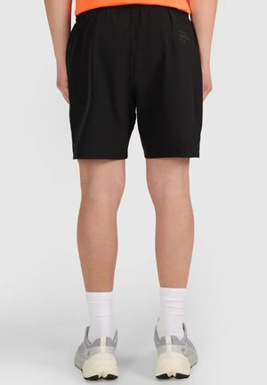 Persona que lleva pantalones cortos negros, calcetines blancos, zapatillas grises y una camiseta naranja, vista desde atrás sobre un fondo liso.