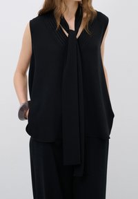 Femme portant un haut noir sans manches avec un long foulard noué au cou et un pantalon noir assorti, main dans la poche, bracelet gris au poignet.