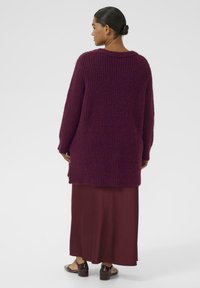 Cardigan oversize in magenta scuro, con texture a coste e scollo ampio, indossato sopra una lunga gonna bordeaux e scarpe basse marroni.