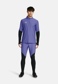 Lila Sportshirt mit halbem Reißverschluss und langen schwarzen Ärmeln, kombiniert mit lila Shorts und schwarzen Leggings, die Logos und ein elegantes Design aufweisen.
