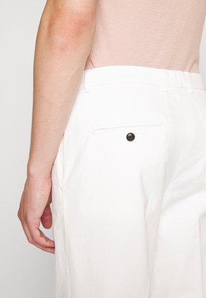 Close-up van een persoon die witte broek draagt met een knoop op de achterzak en een lichtroze mouwloos topje, waarbij de arm en de zijtaille zichtbaar zijn.